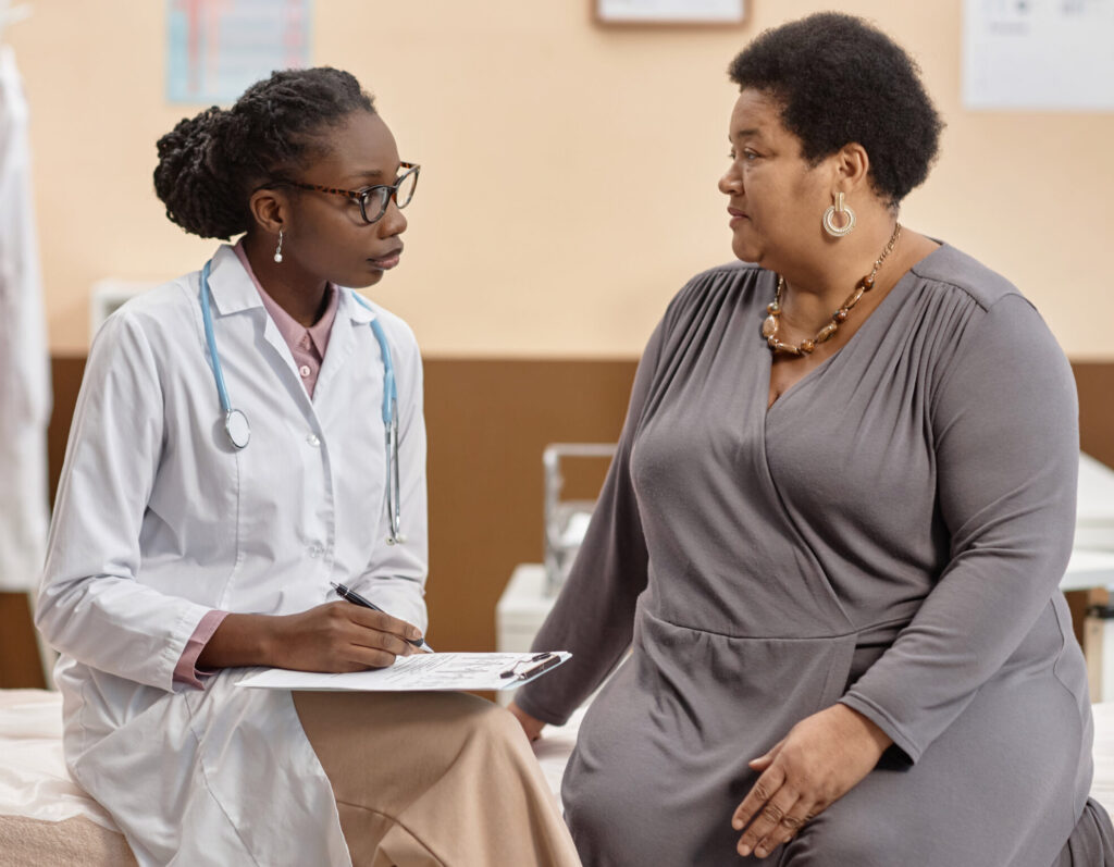 Medium full shot African American woman physician sitting on examination table holding clipboard while listening to mature female patients complaints